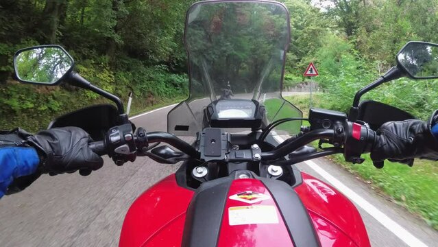POV Biker rides on a motorbike by forested mountain road in Italy. Motorcyclist on motorcycle goes between mountain green trees by landscape highway. Steering wheel view. First-person view. Moto trip
