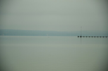 A long lonely footbridge reaches into the foggy Ammersee in Bavaria. Mountain ranges on the horizon
