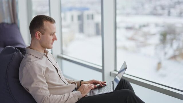 Calm man in bean bag chair works on laptop. Male sitting in front of panoramic window in spacious office. Blurred backdrop.