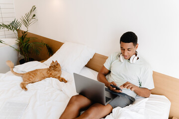 Black man using cellphone and laptop while sitting with his cat on bad