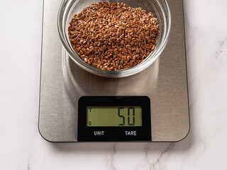 Raw flax seeds in a glass bowl on a digital kitchen scale. 50g flax seed on the scales displaying...