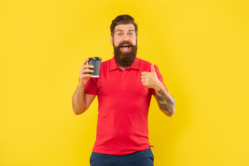 Excited man in casual tshirt giving thumb holding disposable cup yellow background, paper-cup coffee