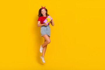 Fototapeta na wymiar Full length photo of sweet small girl jump with present wear t-shirt hairband skirt shoes isolated on yellow background