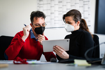 Handsome businessman and businesswoman working together in office during coronavirus pandemic. Two coworkers wearing white and black color FFP2 face masks while analyzing data on tablet screen.