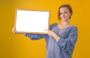 attractive woman smiles and points a finger at a blank white board in her hands on a yellow background, mockup