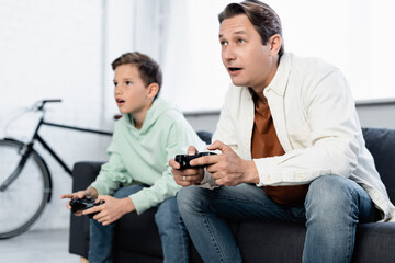 Fototapeta premium KYIV, UKRAINE - OCTOBER 11, 2021: Man playing video game with preteen son on couch at home