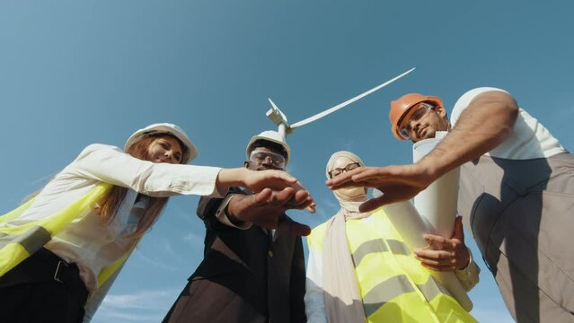 Men and women in safety helmets having successful meeting outdoors. Group of four multicultural partners stacking hands together and smiling on camera while standing among wind turbines