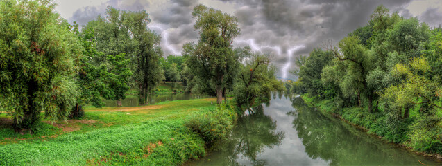 Naturlandschaft im Grünen mit Bach, Bäumen und saftigen Wiesen bei einem aufziehenden...