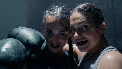 Portrait Shot of Two Female Kick Boxers Hugging Each Other, Wearing Dental Guards and Black Boxing Gloves As They Look Straight At The Camera Smiling. Strong and Confident Women Boxers Training. - Powered by Adobe