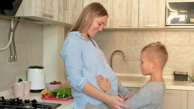 Pregnant mom with kid playing together and huging in the kitchen. Son kissing mom's pregnant belly