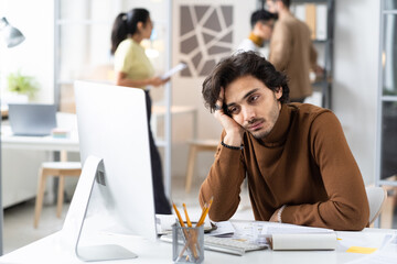 Young businessman sitting at his workplace and looking at computer monitor with bored expression, he tired of his work