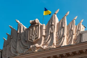 Gordijnen The national flag of the state of Ukraine flutters in the wind on top of the facade of an old building Kharkiv ODA. Yellow-blue flag. National emblem of Ukraine. Ukraine under attack. © Andrii A