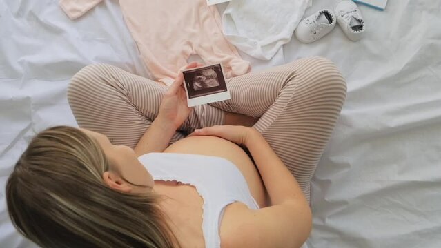 Top view of pregnant woman in the bed preparing baby clothes at home and looking at the ultrasound picture baby, pregnancy and birth concept