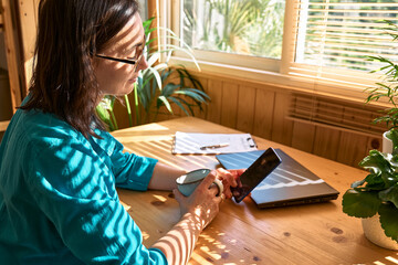 Woman wearing eyeglasses sitting by window and using smartphone checking email news online, looking for friends in internet social networks, writing blog or made call by work. Freelance working.