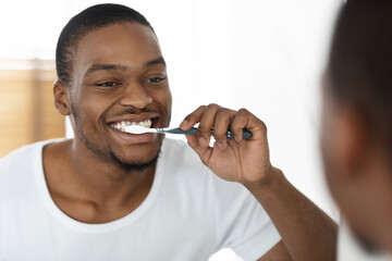 Morning Routine Concept. Happy Black Man Brushing Teeth Near Mirror In Bathroom,