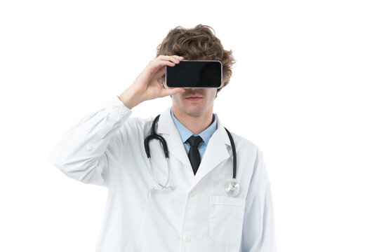 Young doctor in gown suit with stethoscope holding smartphone over foreface to show blank screen.