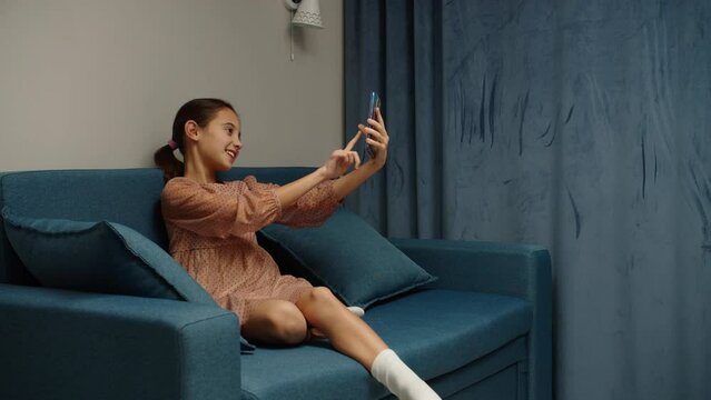 Charming preteen girl taking selfie, using mobile phone, photo messaging while sitting on couch at home. Lovely female child photographing for social media, using mobile app via smartphone indoors