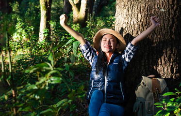 A happy female tourist sitting and raising her hands while lean on tree in the autumn forest with backpack.  looking at the landscape, backpack next to her, Holiday and vacation.