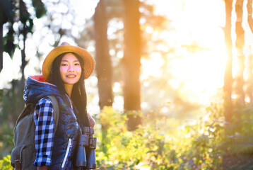 Asian young beautiful female tourist wearing hat, backpacking, holding binoculars, smiling with happiness for traveling at sunset in nature. Outdoor holiday, vacation and travel at the golden hour.