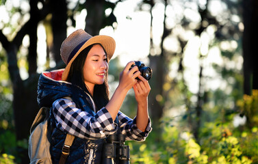 Asian young beautiful female tourist wearing hat, backpacking, holding camera, smiling with happiness for traveling at sunset in nature. Outdoor holiday, vacation and travel at the golden hour.