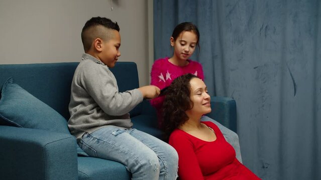 Lovely multiethnic kids combing hair to mother, doing her hair at home. Cheerful woman relaxing, enjoying pastime with her diverse mixed race son and daughter indoors. Family with two preteen children