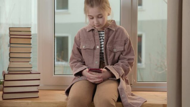 girl sits on the windowsill near the books and looks at the phone