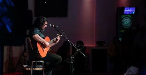 Singer-songwriter plays guitar and sings at acoustic concert in a club with an intimate atmosphere. Silhouettes of the public in the background © MiguelÁngel Olivares