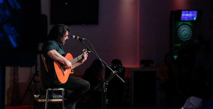 Singer-songwriter plays guitar and sings at acoustic concert in a club with an intimate atmosphere. Silhouettes of the public in the background