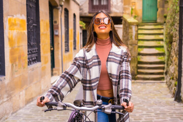 Fototapeta na wymiar Pretty latin woman with sunglasses doing bicycle sightseeing in the city through the old town. Eco tourism in spring on vacation, model smiling