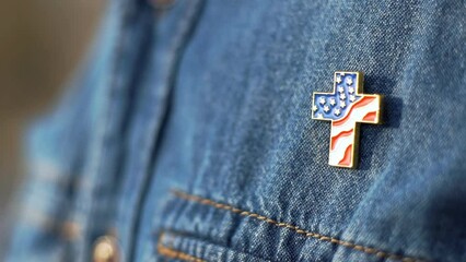 Close up of Christian cross pin with American flag colors is pinned on blue jeans jacket. Patriotism and religious rights concept. - Powered by Adobe