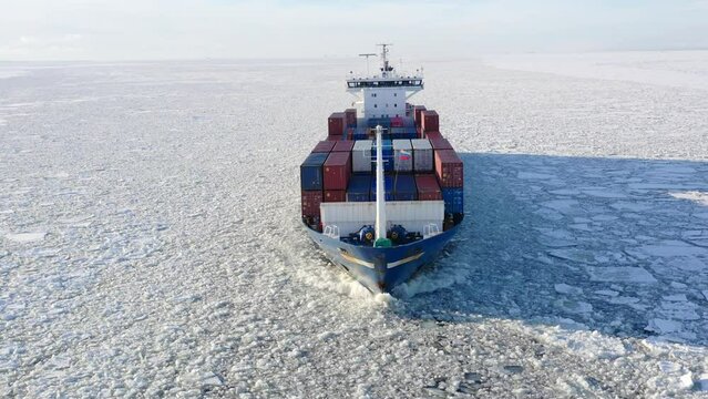St. Petersburg, Russia, winter 2022: aerial view Cargo container ship running through the ice floe, front side of the ship. Ice floes brakes on side of ship