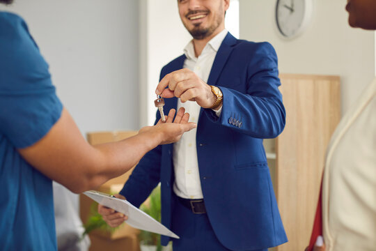 Male home salesman, real estate agent or realtor gives keys to new home to young married couple. Cropped image of man in formal suit handing over keys to new owners of apartments. Real estate concept.