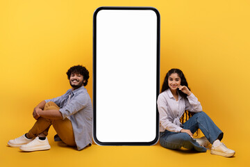 Excited indian couple sitting near giant smartphone with blank white screen, demonstrating copy...