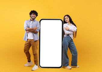 Young indian spouses posing near huge smartphone with mockup, promoting mobile app or website, free space
