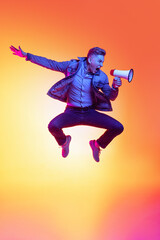 Fototapeta na wymiar Full-length portrait of young excited man jumping with megaphone isolated on orange background in neon light, filter. Concept of emotions, beauty, fashion