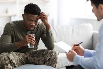 Emotional black military man drinking water while talking to psychotherapist