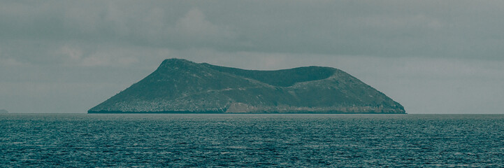 Isolated island in the Galapagos, Ecuador, surrounded by the clear blue waters of the Pacific Ocean under a cloudy sky.