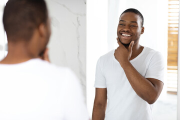 Happy Young Black Man Looking In Mirror In Bathroom And Touching Face