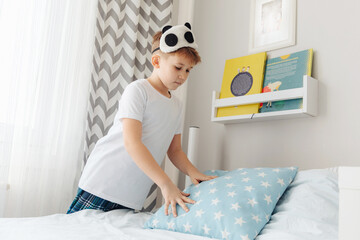 Young boy making his bed in the morning in sunny bedroom.