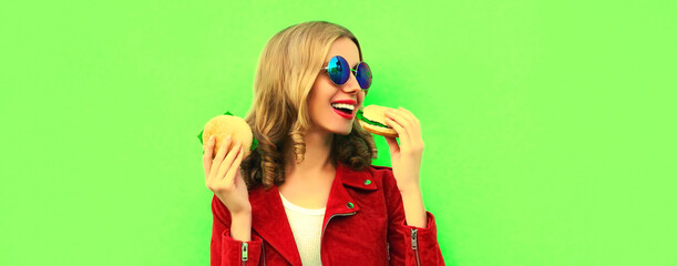 Portrait of stylish happy smiling young woman eating tasty big burger fast food on green colorful background