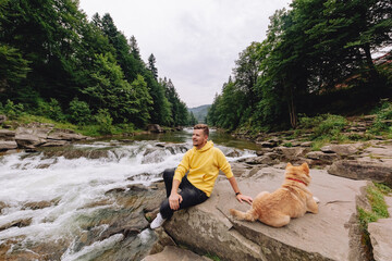 A man relaxes near a mountain river while sitting on a rock. A brown dog is running nearby.