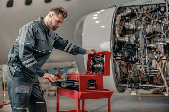 Bearded man airplane maintenance technician in work uniform checking box with instruments while repairing plane