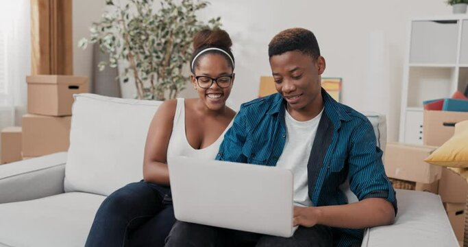 A married couple sits on the couch in their new apartment after moving in, the man holds laptop in his lap, they search together for furniture, furnishings, decorations on websites