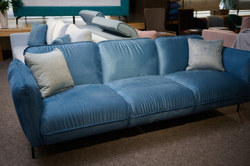 Beautiful minimalist stylish blue velour settee with light gray cushions, displayed on sale in the furniture store showroom. Exhibition of soft furnishing in the furniture store showroom