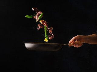 Octopuses in a frying pan with vegetables in a frozen flight in the chef's hand on a black background. Restaurant cuisine recipes. Cookbook, food blog, restaurant, hotel, advertising.