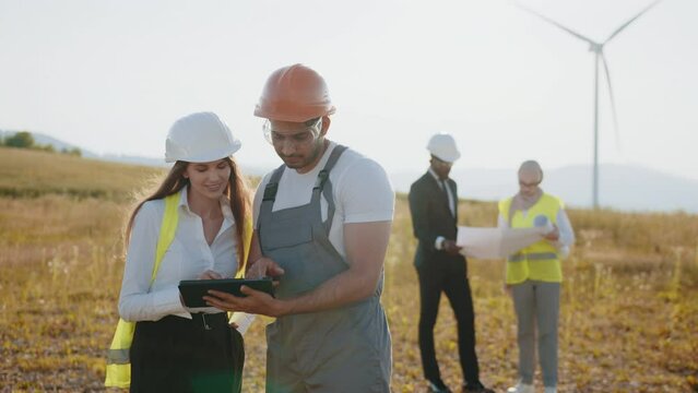 Four colleagues using blueprints and tablet outdoors. Multiracial colleagues controlling work of windmill farm. Eco for electric power. Engineers explores future wind turbine installation projects