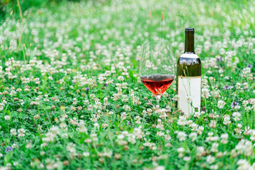 Classic opened bottle of wine standing near big wine glass filled with some pink, red, portwein or madeira wine on grass field or meadow with white wildflowers on summer day outside. Alcohol drink