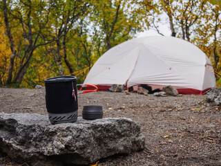 A pot for a gas burner with a mug sits on a stone against the background of a tent.
