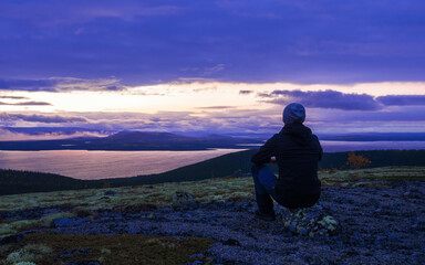 A lone tourist in a hat sits on a stone in the mountains against the backdrop of a lake at sunset, view from the back.