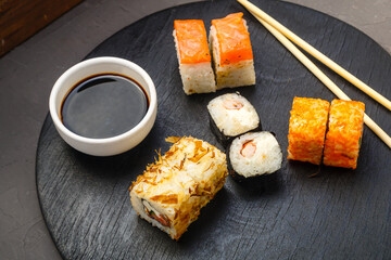 Rolls with fish and shrimp on a round board next to sticks, soy sauce on a dark background.